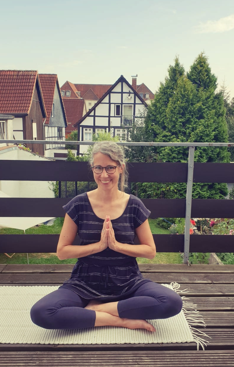 Katja sitzt im Schneidersitz auf einer Terrasse, die Hände vor der Brust